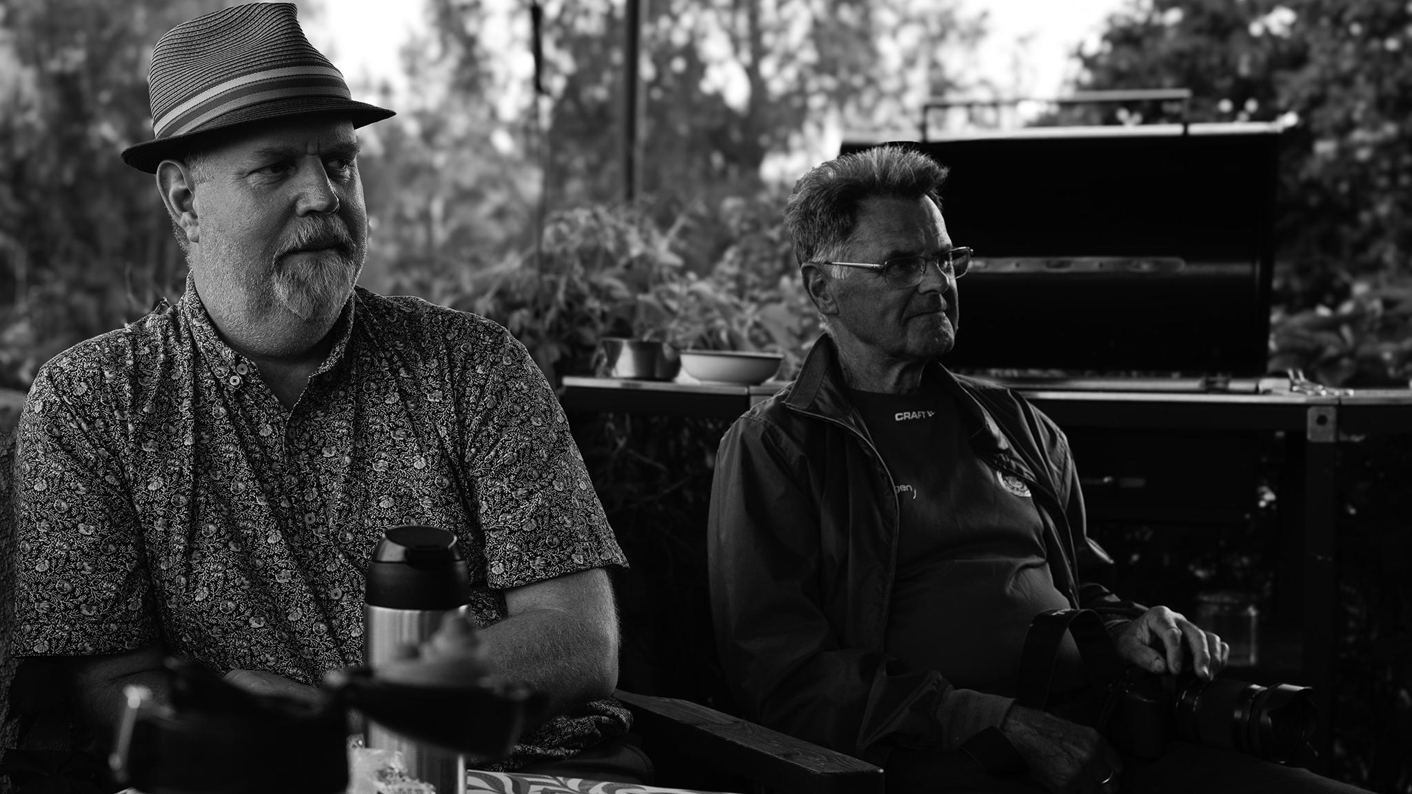 Two elderly men sit outdoors, one with a hat and the other with glasses; plants and a grill in background.