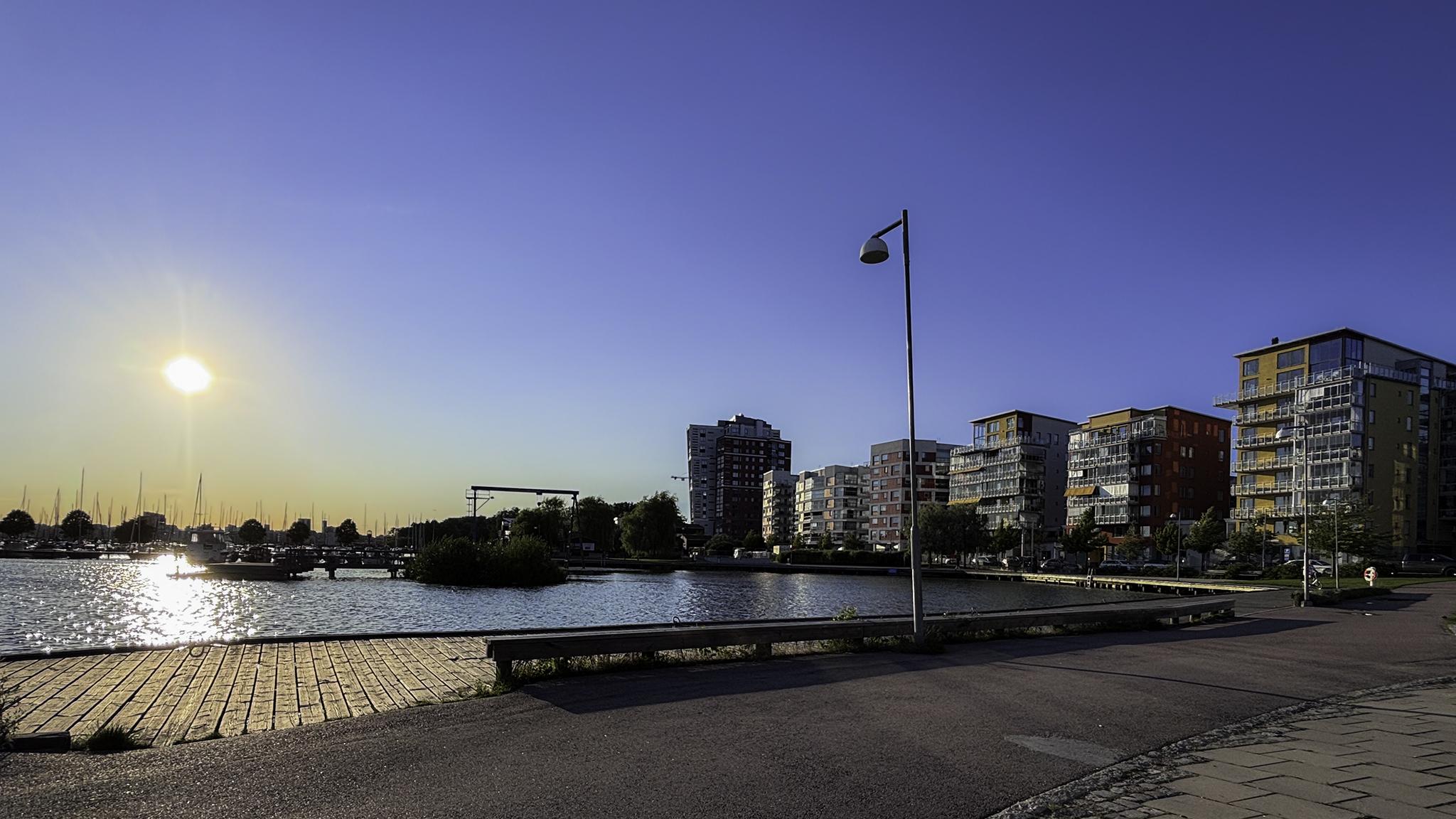 Sjö med segelbåtar i solnedgång, moderna höghus till höger och grönska längs strandpromenaden.