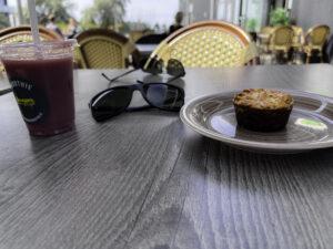 Plastmugg med smoothie, svarta solglasögon, tallrik med bakelse på bord utomhus, möbler i bakgrunden.