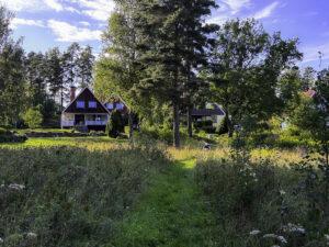 Tvåvåningshus med bruna och vita fasader i en trädgård omgiven av träd och grönska, klarblå himmel.