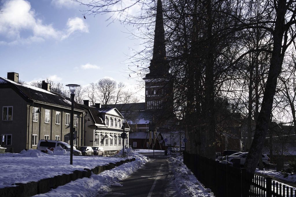 Vinterbild av en gata med gamla hus och en kyrka med hög spira i bakgrunden. Snö ligger längs vägen och kala träd reser sig mot en blå himmel.