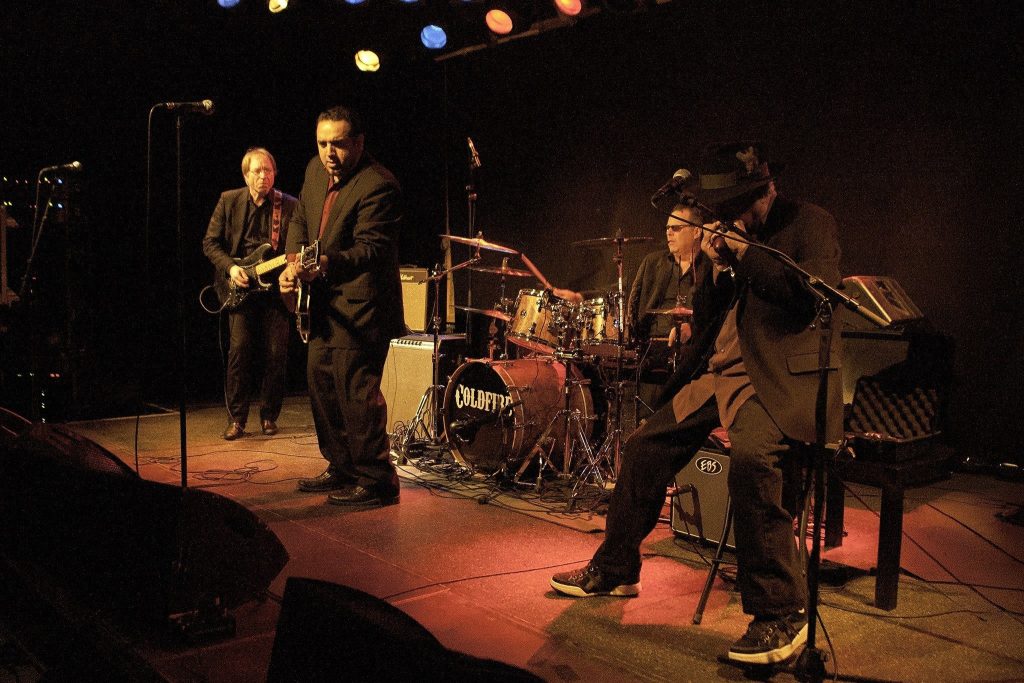 Konsertbild av ett bluesband på scen: en basist och gitarrist till vänster, en trumslagare i mitten och en pianist till höger, i dämpad scenbelysning.