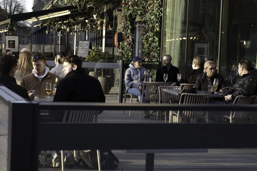Kvällsbild av en uteservering där gäster sitter vid bord utanför en upplyst restaurang med gröna växter på fasaden.