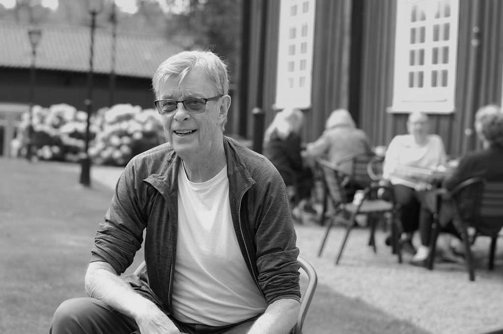 En man sitter utomhus på Café Malin i Västerås och ler, med fler gäster och blommande buskar i den suddiga bakgrunden.