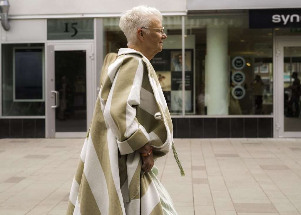 Äldre kvinna med vitt kort hår går på gågata iklädd beige och vit rutig poncho – slow fashion från Global Living av Eva Wells