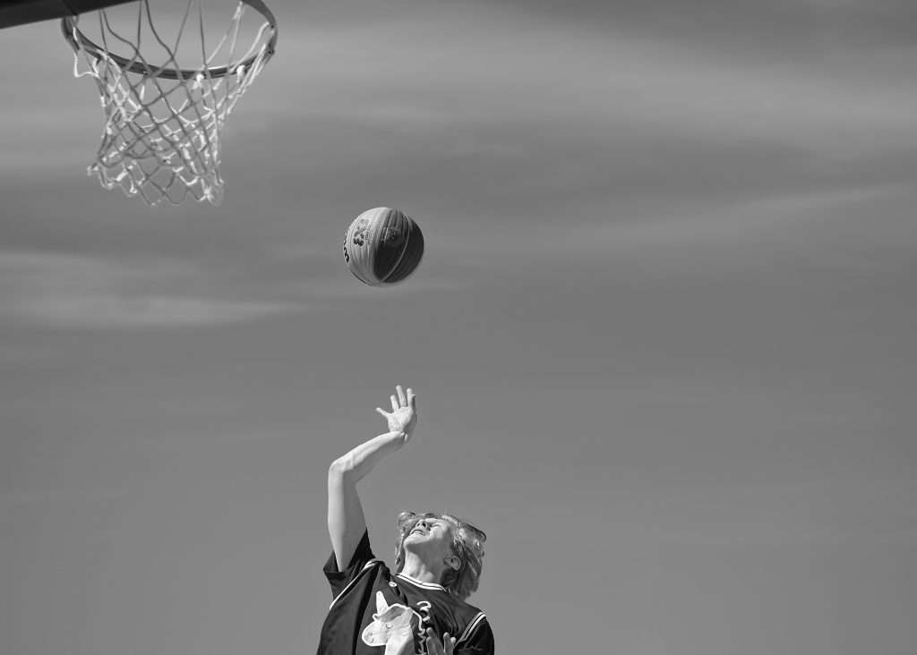 Svartvit bild underifrån av en ung basketspelare med nummer 3 på tröjan som skjuter bollen mot korgen mot en molnig himmel, med korgen synlig i övre vänstra hörnet.