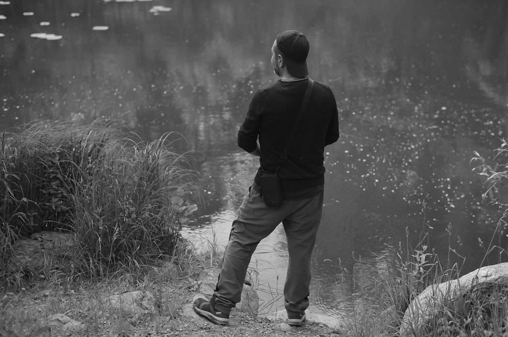 Svartvitt fotografi tagit bakifrån på en man i keps, tröja och friluftsbyxor som står vid en strandkant och blickar ut över ett lugnt, speglande vatten. Högt gräs kantar vattnet i förgrunden.