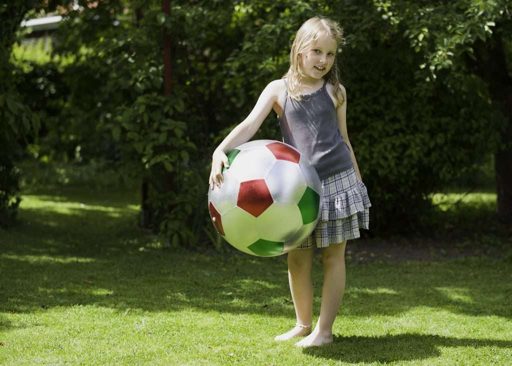 Florence står barfota på en gräsmatta i sommarträdgården och håller en stor färgglad fotboll, omgiven av grönska.
