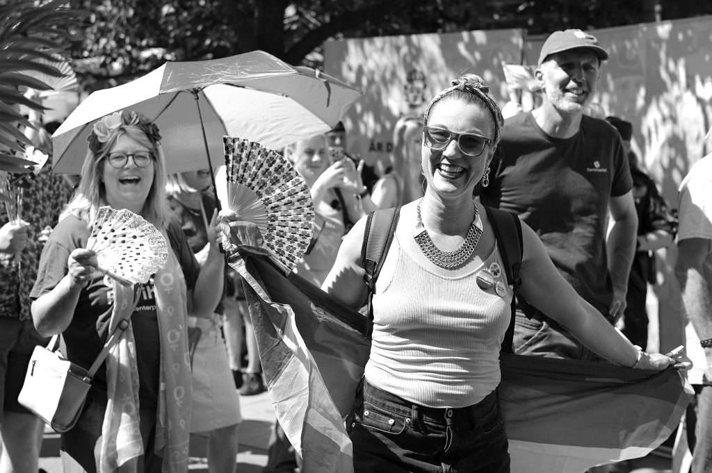 Svartvit bild från ett Pride-tåg en solig dag. Glada människor går framåt – en kvinna i tanktopp och solglasögon ler stort och bär en regnbågsflagga, en annan kvinna med blomsterkrans skrattar under ett parasoll och håller en solfjäder. I bakgrunden går fler deltagare, flera med t-shirts från Centerpartiet.