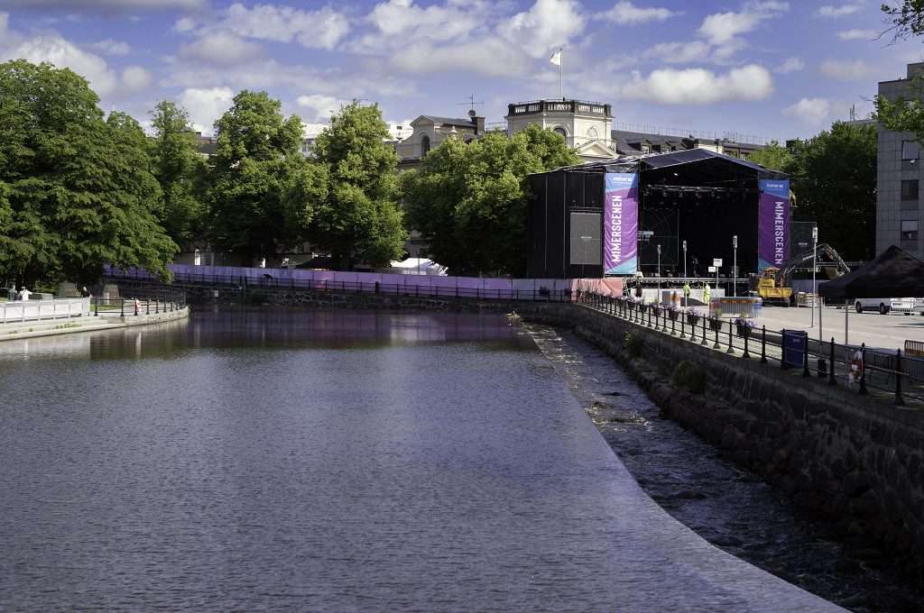 Mimerscenen rest vid Svartån i Västerås inför Cityfestivalen, med scenrigg och banderoller längs ån en solig sommardag