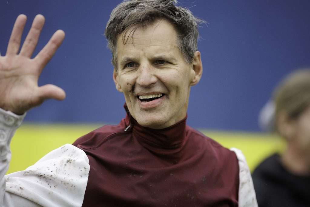 Leende jockey i vinröd och vit ridställning vinkar med handen efter segern i Grand National på Strömsholm
