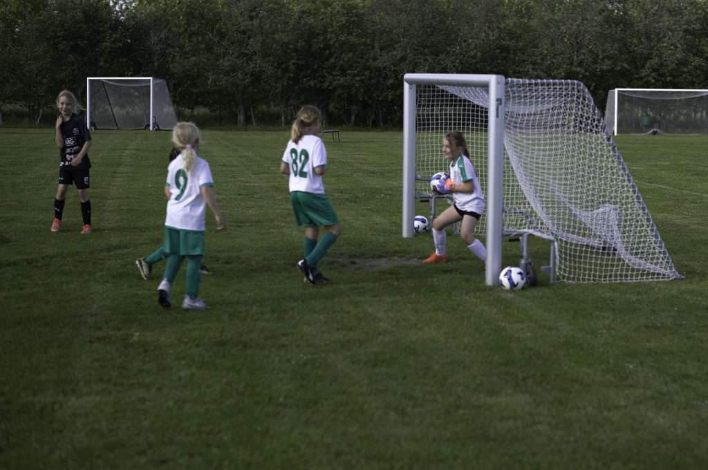 Fyra barn i Tillberga IK:s vita och gröna fotbollsställ spelar på en gräsplan en sommarkväll. En ung målvakt vid målet håller bollen i händerna och ler, medan två spelare med nummer 9 och 82 på ryggen springer mot målet och ett barn i svart ställ tittar på från vänster. Flera bollar ligger på planen och i bakgrunden syns träd och fler fotbollsmål.