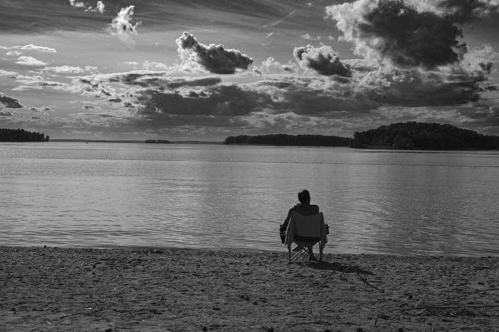 Svartvit bild av en man som sitter ensam i en campingstol på en sandstrand och blickar ut över en stilla sjö. Himlen fylls av dramatiska, böljande moln, och i bakgrunden syns skogklädda uddar och öar vid horisonten.