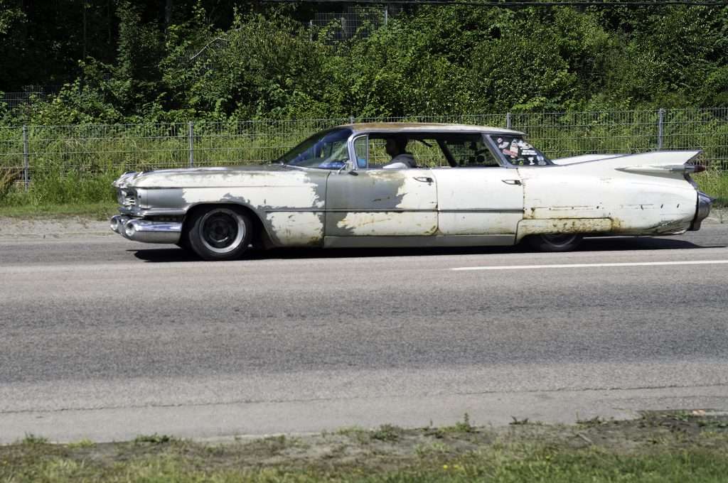 Färgbild av en vit klassisk Cadillac med rostiga fläckar och avflagnad lack, fotograferad i profil på en väg. Bilen har karakteristiska bakre stjärtfenor och körs av en man, med grönska och ett staket i bakgrunden.