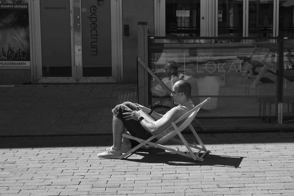Svartvit gatubild av en man som sitter tillbakalutad i en solstol på ett stenlagt torg och läser en bok. Han bär shorts, t-shirt och glasögon. Bakom honom en glasfasad märkt "expectrum" och "bar Lokal", och hans spegelbild syns i glaset.
