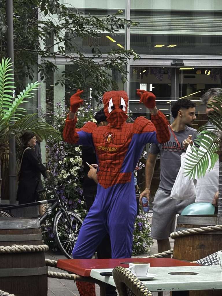 Färgfoto från en uteservering i stadsmiljö. En person i röd och blå spindelmansdräkt med texten "Spider Bar" på bröstet står med båda händerna uppsträckta i webbslingar-pose. Runt omkring passerar förbipasserande, och i förgrunden syns cafébord med en kaffekopp, träfat och rep.