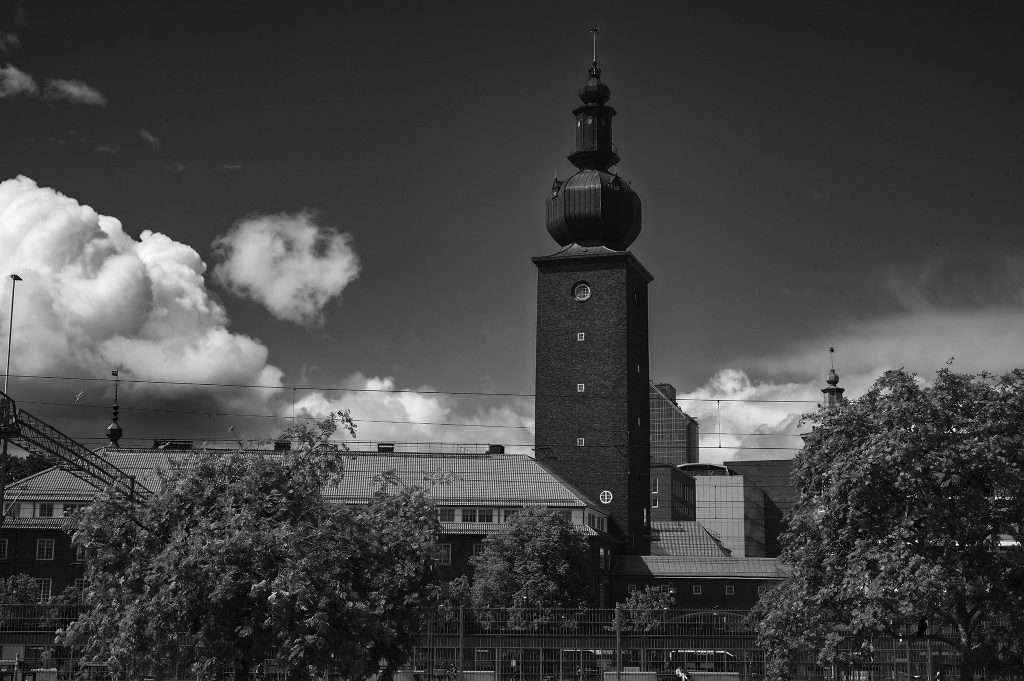 Svartvitt foto av ABB:s svenska huvudkontor – ett högt tegeltorn med kupolformad lökspira som reser sig över kontorsbyggnaderna bakom lummiga träd och ett järnstaket, mot en himmel med stora moln.