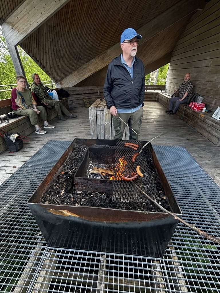 En man i keps och blå jacka grillar korv över öppen eld i en järngrill inne i vindskyddet vid Hälleskogsbrännan, medan andra vandrare vilar på bänkarna runt om.