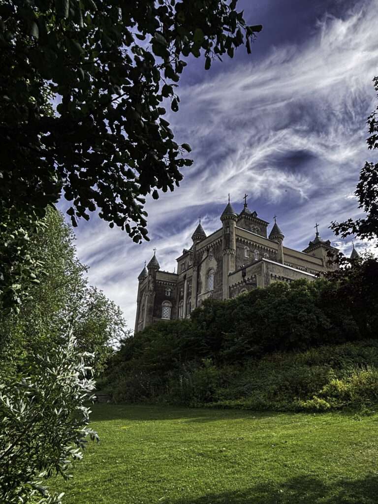 Stora Sundby slott sett underifrån från gräsmattan nedanför slänten. Slottets fasad med torn och gröna kopparspiror reser sig ovanför en tät buskridå, mot