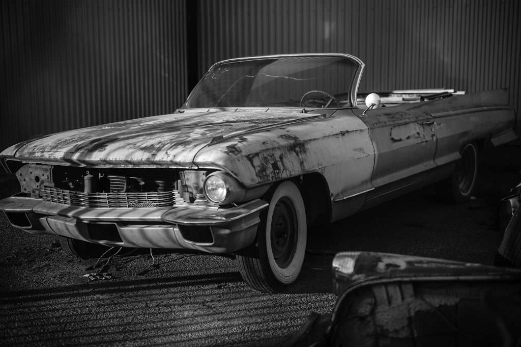 Svartvitt foto av en gammal Cadillac-cabriolet från tidigt 1960-tal, parkerad framför en korrugerad plåtvägg. Bilen är rostig och sliten, saknar en strålkastare och har vitsidiga däck. En spräckt vindruta och renoveringsdetaljer syns, och lösa karossdelar ligger på marken framför.