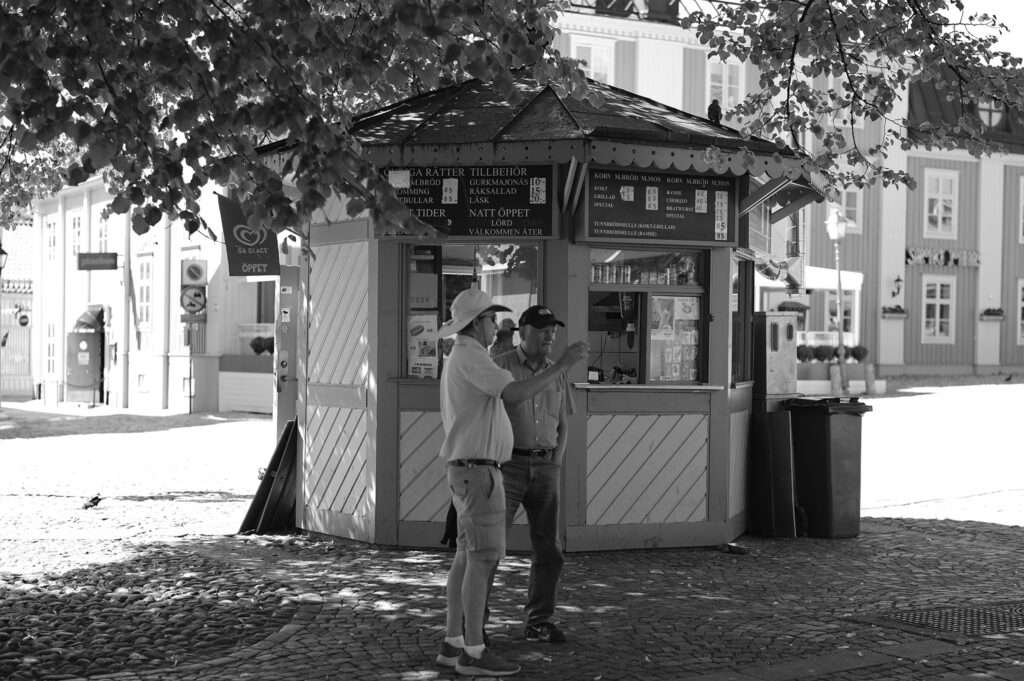 Två män samtalar framför en gammaldags korvkiosk på ett kullerstenstorg.