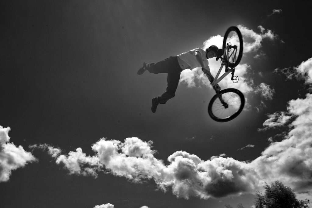 En cyklist svävar högt i luften med kroppen utsträckt vid sidan av cykeln, fotograferad underifrån i motljus mot en himmel med vita moln.