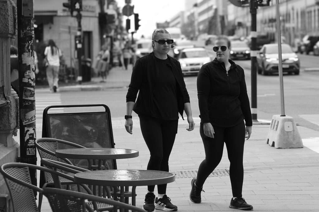 Svartvit gatubild av två kvinnor i svarta träningskläder och solglasögon som går i rask takt längs en trottoar. Till vänster står tomma caféstolar och runda bord vid en affischklistrad stolpe. I bakgrunden gatukorsning med bilar och fotgängare.