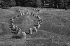 Svartvit bild av en skeppssättning, resta stenar placerade i en lång oval skeppsform på en gräsbevuxen slänt. Stenarna kastar tydliga skuggor i solljuset. Längst bort syns ytterligare stenar i samma formation och en rad lövträd.