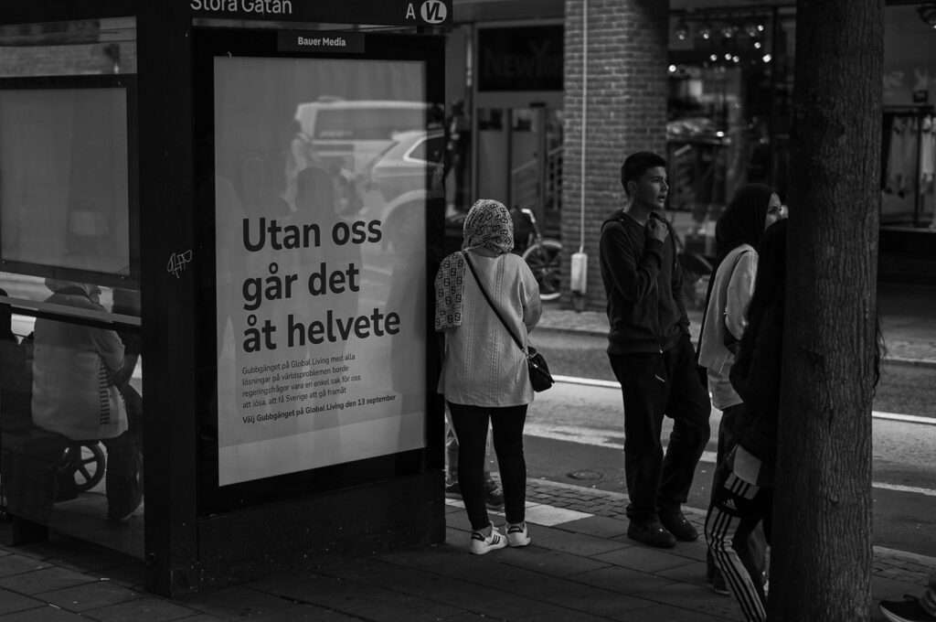 Busskur på Stora Gatan med en stor affisch med texten "Utan oss går det åt helvete" och Gubbgänget på Global Living som avsändare, medan några personer väntar på bussen bredvid.