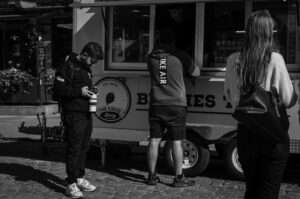Foodtruck på kullerstenstorg i solljus, en man beställer i luckan medan en fotograf med kamera och en kvinna med väska står bredvid.