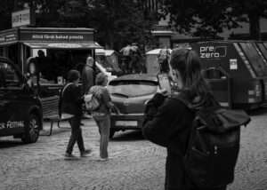 Kvinna med ryggsäck fotograferar med mobilen på ett kullerstenstorg där en foodtruck med skylten "Alltid färsk bakad potatis" står, och en bronsskulptur av cyklister syns bakom bilarna.