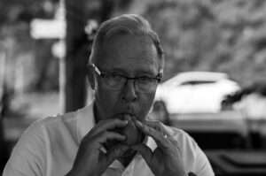 Svartvitt porträtt av Stefan, en man med grått bakåtkammat hår, glasögon och vit pikétröja. Han blåser i en liten ocarina som han håller med båda händerna framför munnen och tittar koncentrerat in i kameran. I den suddiga bakgrunden syns träd och en vit bil.