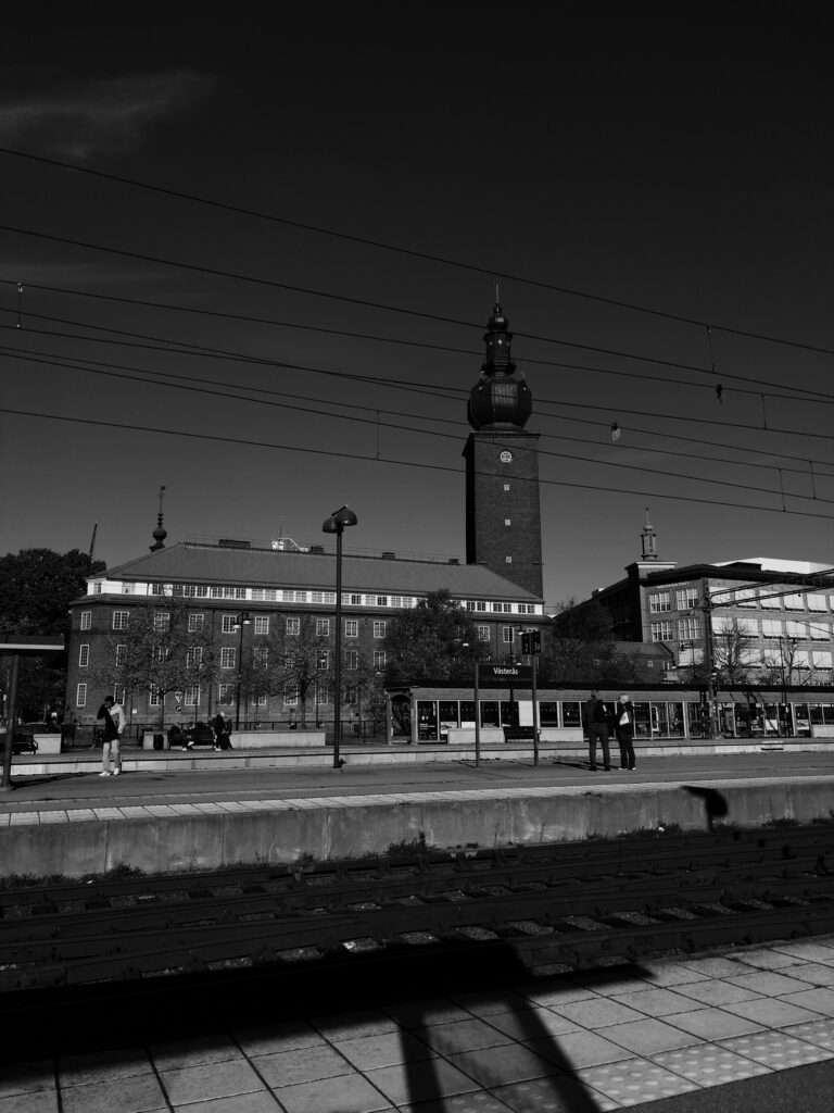 Svartvitt foto från Västerås centralstation, taget över spåren mot en perron där några personer väntar. En skylt med texten “Västerås” står på perrongen. Bakom ligger en stor tegelbyggnad med ett högt torn som har en klocka och en lökformad kupol – det gamla ASEA-tornet, i dag en del av ABB:s huvudkontor. Kontaktledningar korsar den mörka himlen och i förgrunden syns skuggor på perrongplattorna.