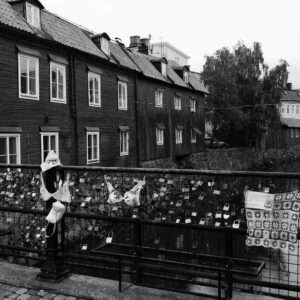Svartvitt foto från ett broräcke över Svartån, fullt av kärlekslås i alla storlekar. På räcket hänger också två behåar, en liten vit handväska och en virkad mössa. Till höger sitter en virkad filt med ett bandsymbol och en skylt med QR-kod. Bakom ån står gamla timmerhus med vita fönster och ett hängande träd vid stenkanten.