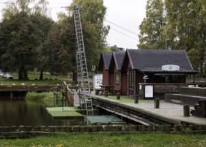 Wakeboardanläggning vid vattnet en mulen höstdag. En hög aluminiummast med stegpinnar bär kabelbanan över dammen. Bredvid ligger startbryggor klädda med grön konstgräsmatta, en träbrygga och tre röda bodar med svarta tak. På en av dem sitter en wakeboard med texten Cable Bar. Bryggan är tom, och bakom bodarna står träd i höstfärger.