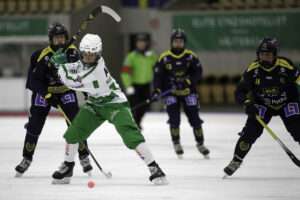 Galina Mikhaylova i Västerås SK:s grönvita dräkt lyfter klubban högt för ett slag mot den röda bollen framför sig på isen. Runt henne står tre Skirö-spelare i mörkblå och gul dräkt, och i bakgrunden syns en domare i grön tröja och läktarstolar.