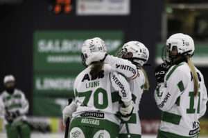 Tre spelare i Västerås SK:s grönvita dräkt firar tillsammans, sedda bakifrån. Nummer 70 med namnet Ljung på ryggen kramas om av nummer 18, som bär kaptensbindel, medan nummer 11 står bredvid. I bakgrunden syns en oskarp lagkamrat och gröna reklamskyltar.